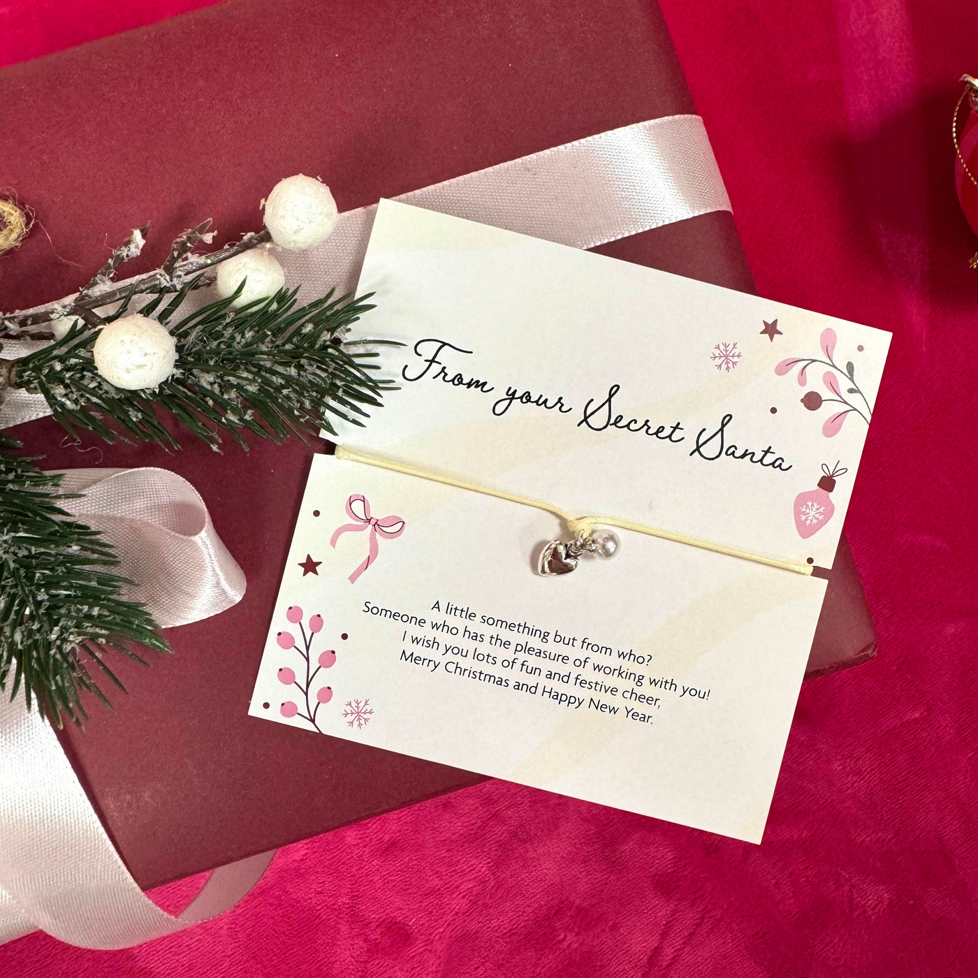 A small silver heart & pearl charm attached to a cream card that reads 'From your Secret Santa,' styled on top of a dark red gift box with a white ribbon and holiday greenery on a red fabric background.