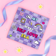 Purple chequered card reading "In my self love era" with a beaded friendship bracelet with the word "LOVE"