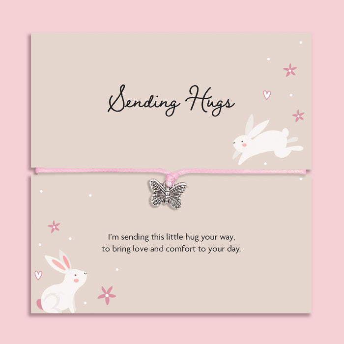 Pink string bracelet with a silver butterfly charm, displayed on a cream card with bunny illustrations and the message “Sending Hugs – I’m sending this little hug your way, to bring love and comfort to your day.