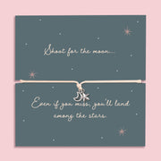 Flat lay image of a "Shoot for the Moon" charm bracelet displayed on a navy starry-themed card with the inspirational quote "Shoot for the moon... Even if you miss, you’ll land among the stars." The bracelet features silver moon and star charms on a soft cream cord, designed as an uplifting gift for women.