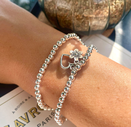 A women's wrist with two silver-plated beaded bracelets resting on a table. The charms on the bracelets are a flower and hollow heart.