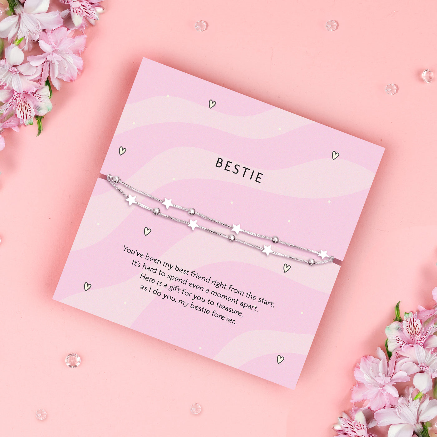 Silver-plated star bracelet on a pink wavy poem card with a meaningful sentiment for a friend. Making a perfect gift for a best friend on a pink background surrounded by gems and pink floral flowers.