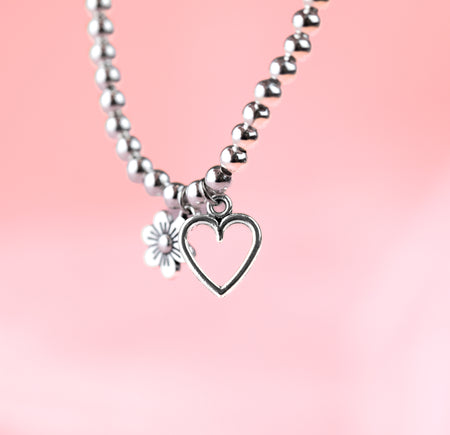 A pale pink background with a dangling beaded bracelet with heart and flower charms. The heart is the focal point with the flower blurred in the background.
