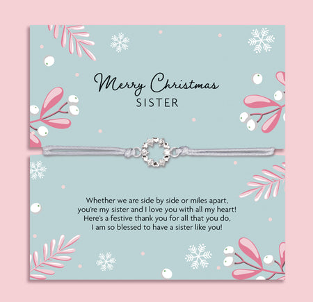 Merry Christmas Sister bracelet featuring a silver wreath charm on an adjustable cord, presented on a festive green card with snowflakes, mistletoe, and pink leaves. Includes a soft white pouch for gifting and safekeeping. A sentimental Christmas jewellery gift for sisters, perfect as a stocking filler.