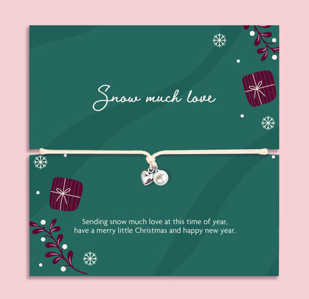 A delicate silver bracelet with a small pearl and silver heart charm is displayed on a festive dark green card. The card features Christmas illustrations, including snowflakes, wrapped gifts, and evergreen branches, along with the text 'Snow much love' and a holiday message.