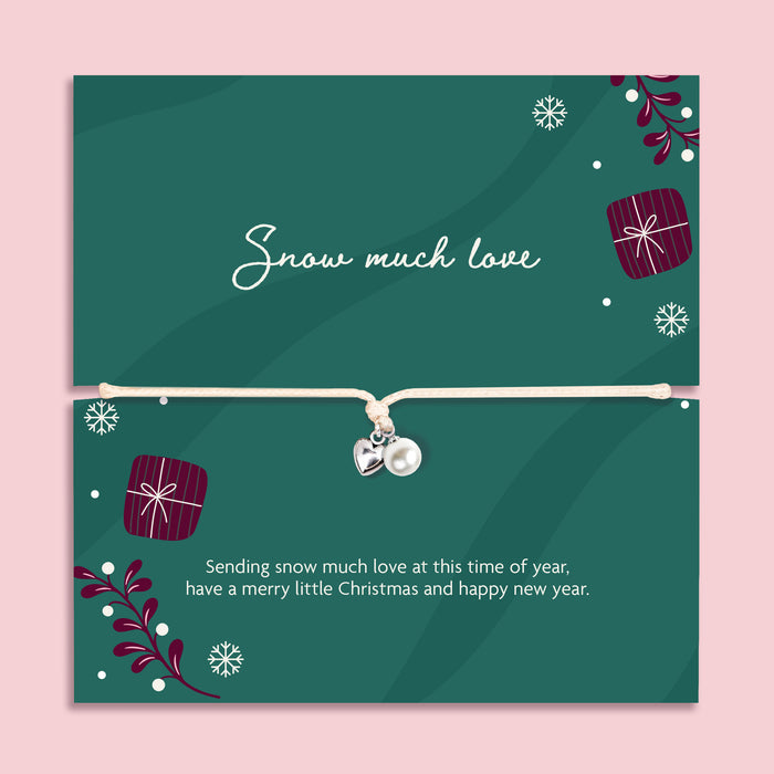 A delicate silver bracelet with a small pearl and silver heart charm is displayed on a festive dark green card. The card features Christmas illustrations, including snowflakes, wrapped gifts, and evergreen branches, along with the text 'Snow much love' and a holiday message.