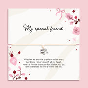 A friendship bracelet featuring a cream card with elegant black script reading 'My special friend' and a matching poem on the bottom, with a bracelet with a small silver heart charm and bow. The cards are decorated with pink and burgundy floral illustrations, set against a solid pink background.