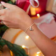 A delicate silver bracelet with a small star and heart charm worn on a person's wrist, shown in a festive holiday setting with wrapped gifts and warm string lights in the background.
