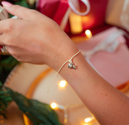 A delicate silver bracelet with a small star and heart charm worn on a person's wrist, shown in a festive holiday setting with wrapped gifts and warm string lights in the background.