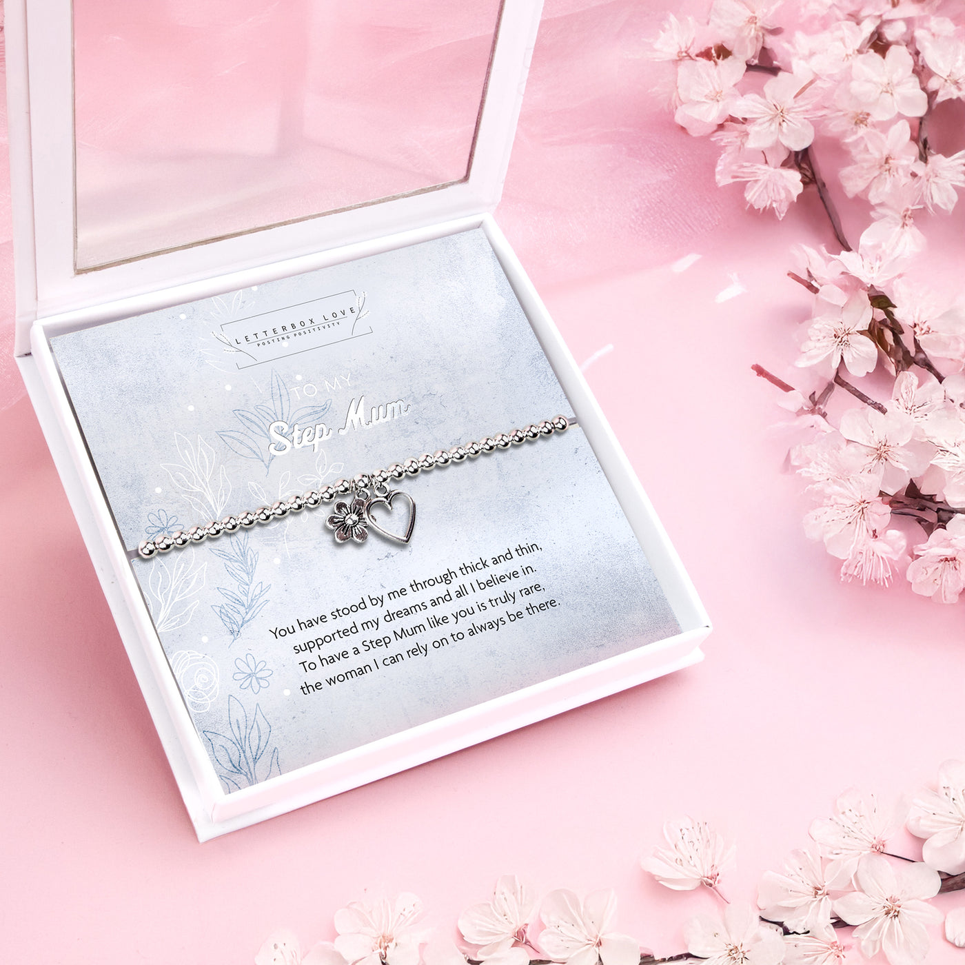 A silver-plated bracelet with a heart and flower charm, presented in a luxury white box with a message, on a pink background with cherry blossoms.