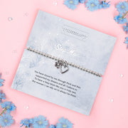 A pale pink background with blue flowers and a blue poem card for a 'step mum'. The bracelet around the card is silver-plated with a heart and flower charm.
