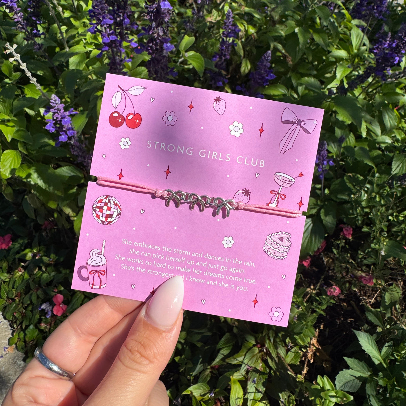 Strong Girls Club pink adjustable cord bracelet displayed on an illustrated empowerment sentiment card, held outdoors in front of bright pink and white flowers.