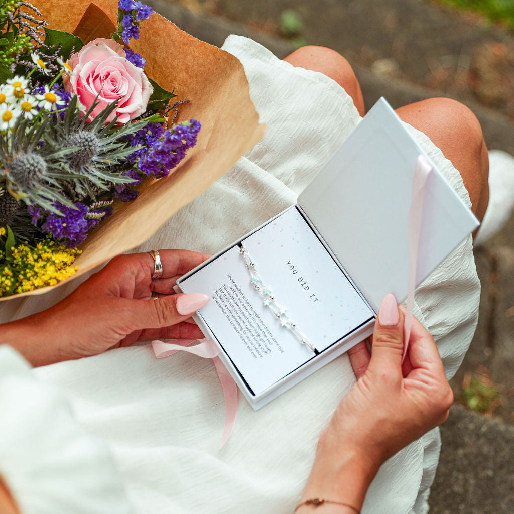 Person holding a bouquet of flowers and a card with text, sitting outdoors. Bracelet - You Did It