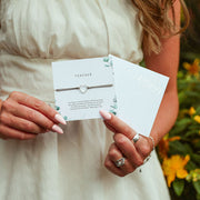 Person holding a Teacher bracelet and its packaging with flowers in the background