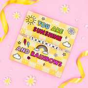 An image of a cheerful motivational card featuring a beaded bracelet with the word "SUNSHINE" spelled out in white letter beads. The card has a bright yellow checkered background with colorful illustrations of a sun, rainbow, lightning bolts, and flowers. Bold text in vibrant colors reads: "You are sunshine and rainbows." The card is decorated with daisies, ribbons, and sparkly accents on a pink background, creating a fun and uplifting vibe. A playful and positive gift designed to bring happiness and light 