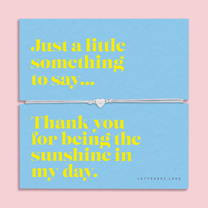 A light blue bracelet card titled "Sunshine" from Letterbox Love, featuring a small heart charm on a grey string bracelet. The card includes the message: "Just a little something to say... Thank you for being the sunshine in my day." The setting is a soft pink background with small yellow flowers and decorative gems scattered around.