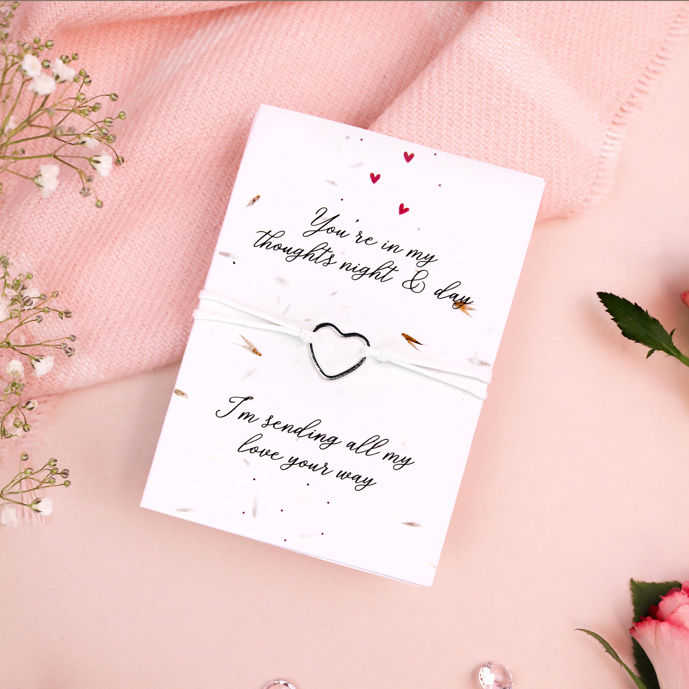 Seeded card with the message 'You're in my thoughts night & day. I'm sending all my love your way' in elegant script. A delicate bracelet with a silver heart charm is attached to the card, which is adorned with tiny red heart illustrations. The card is set against a soft pink background with baby's breath flowers, a pink knitted fabric, and scattered clear gems, creating a warm and comforting aesthetic.