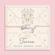 Adjustable peach cord bracelet with a silver 'Taurus' charm and heart pendant, displayed on a zodiac-themed backing card with a bull illustration and traits reading "Resilient, Grounded, Loving".