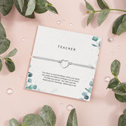 A thoughtful gift card titled "TEACHER" featuring a heartfelt appreciation note for a teacher, accompanied by a simple yet elegant bracelet with a heart-shaped charm. The card's design includes subtle green foliage patterns on a soft grey background, creating a calm and appreciative atmosphere suitable for expressing gratitude to educators.