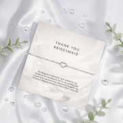 Elegant 'THANK YOU BRIDESMAID' card on a silky white background, featuring a touching gratitude message and a delicate silver bracelet with a heart charm, framed by green leaves. A meaningful bridal party gift.