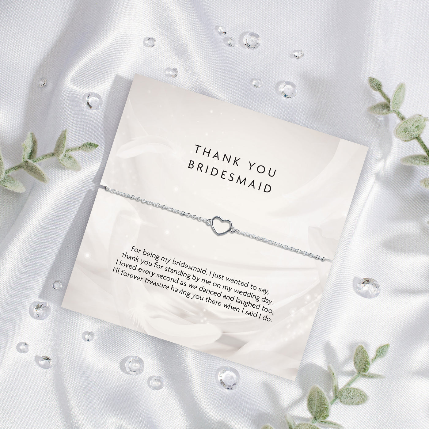 Elegant 'THANK YOU BRIDESMAID' card on a silky white background, featuring a touching gratitude message and a delicate silver bracelet with a heart charm, framed by green leaves. A meaningful bridal party gift.