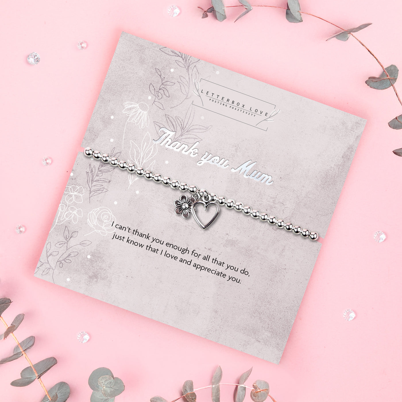 A silver beaded bracelet with a heart and flower charm displayed on a card reading 
