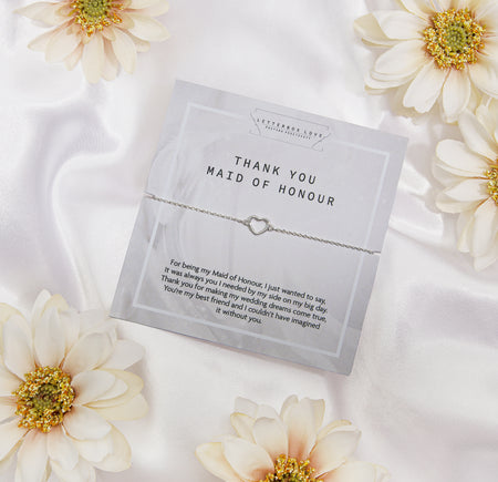 A 'THANK YOU MAID OF HONOUR' appreciation card from Letterbox Love, placed on a silky white fabric background. The card displays a heartfelt message of gratitude and features a delicate silver bracelet with a heart-shaped charm. 