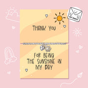 A silver beaded bracelet with a flower charm and a heart charm is displayed on a two-part yellow card that reads 'THANK YOU' and 'FOR BEING THE SUNSHINE IN MY DAY' on a pink background with cute doodles.
