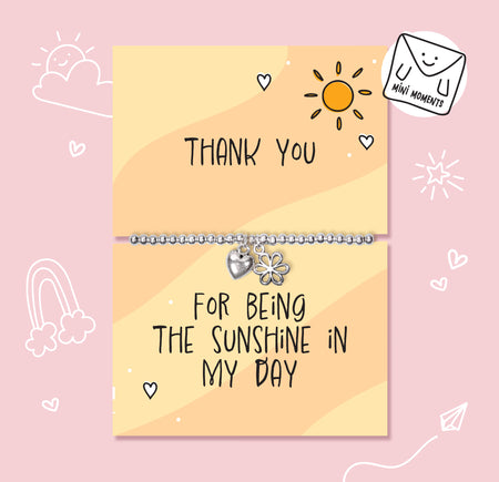A silver beaded bracelet with a flower charm and a heart charm is displayed on a two-part yellow card that reads 'THANK YOU' and 'FOR BEING THE SUNSHINE IN MY DAY' on a pink background with cute doodles.