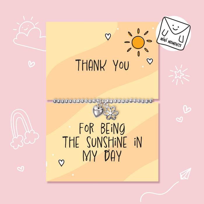 A silver beaded bracelet with a flower charm and a heart charm is displayed on a two-part yellow card that reads 'THANK YOU' and 'FOR BEING THE SUNSHINE IN MY DAY' on a pink background with cute doodles.