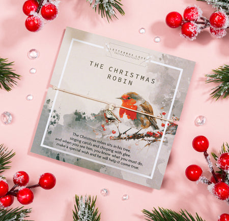 Festive 'The Christmas Robin' card set against a gentle pink backdrop, showcasing a beautifully painted robin amidst snowy branches. The card contains a heartwarming holiday poem and is paired with a dainty bracelet featuring a robin charm.