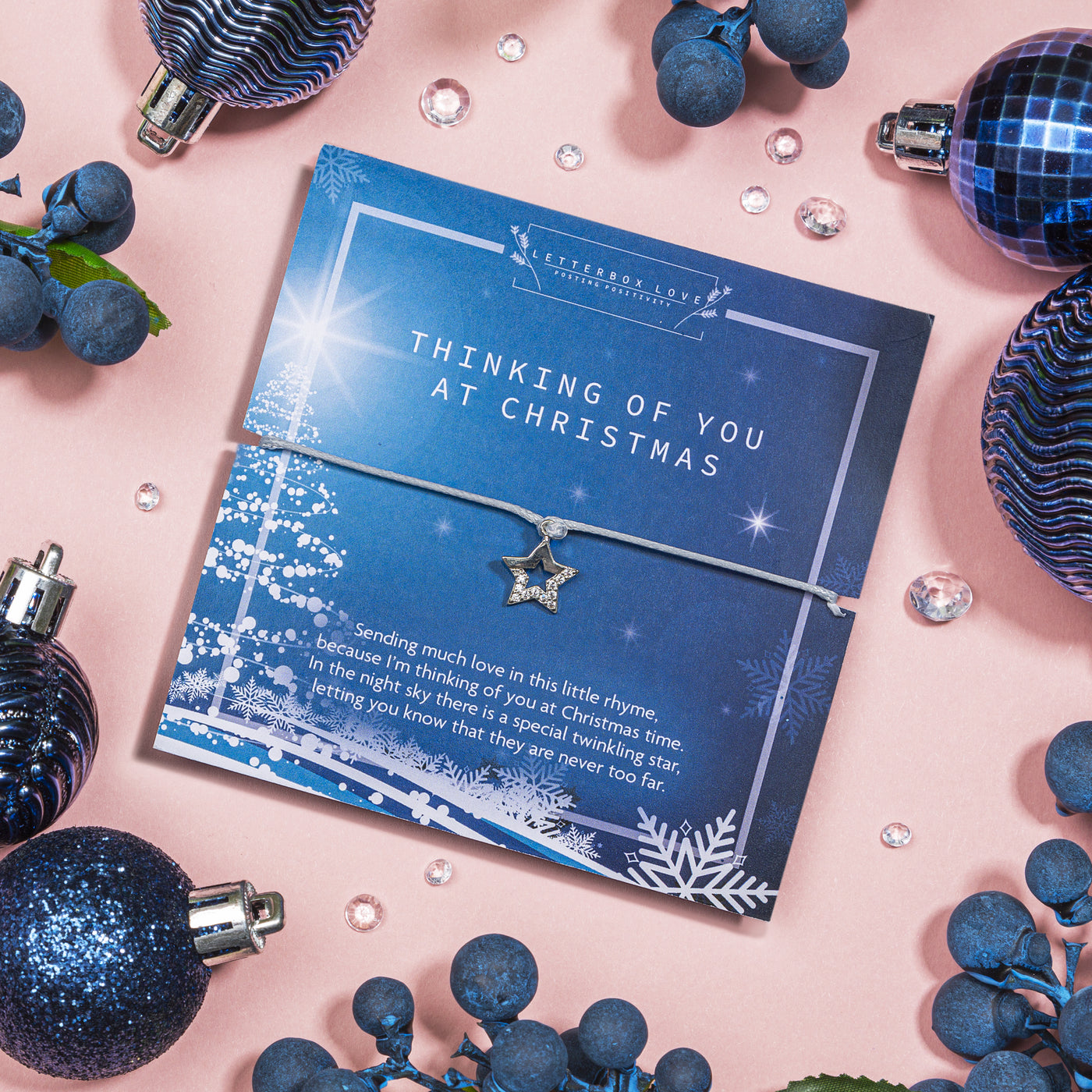 Charming 'Thinking of You at Christmas' card displayed on a pastel pink background. The deep blue card showcases shimmering winter illustrations and a heartfelt message about a special twinkling star. Accompanying the card is a silver star charm bracelet.