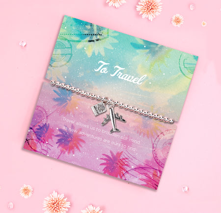 A silver-plated beaded bracelet featuring airplane and passport charms, displayed on a colorful travel-inspired backing card with pastel hues of pink, blue, and purple. The card reads, 