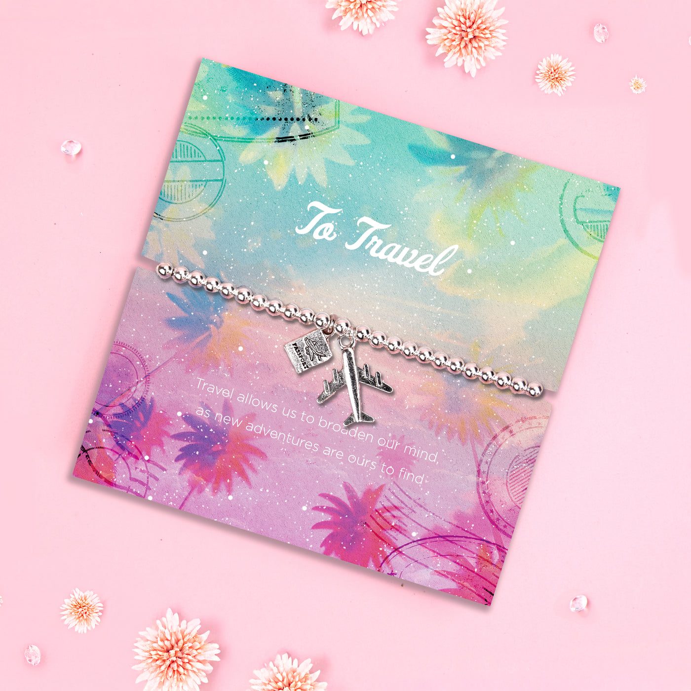 A silver-plated beaded bracelet featuring airplane and passport charms, displayed on a colorful travel-inspired backing card with pastel hues of pink, blue, and purple. The card reads, 