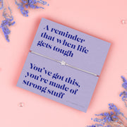 A lavender-colored bracelet card titled "Strong Stuff" from Letterbox Love, featuring a small star charm on a grey string bracelet. The card includes the message: "A reminder that when life gets tough, you’ve got this, you’re made of strong stuff." The setting is a soft pink background with small purple flowers and decorative gems scattered around.