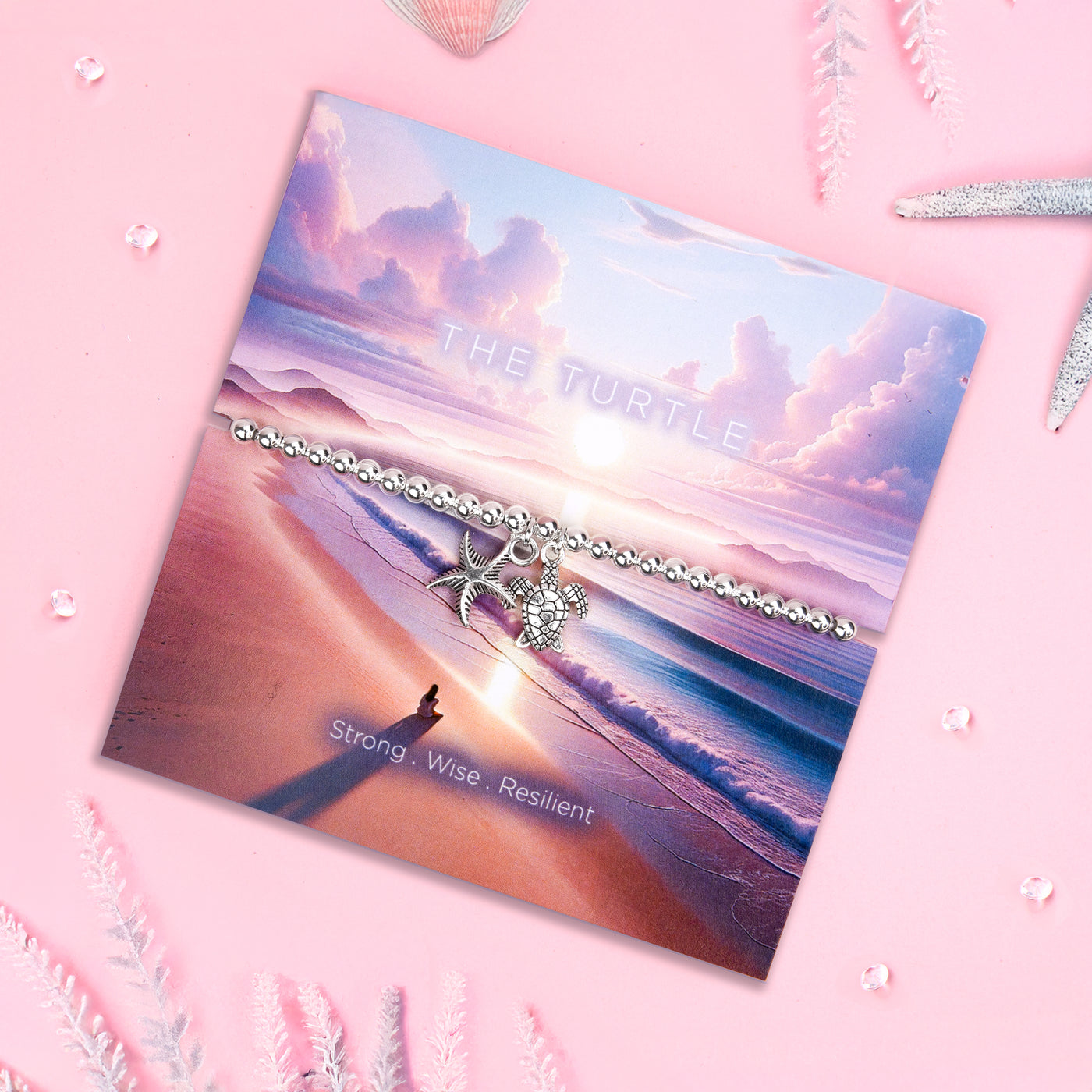 A silver-plated bracelet with a delicate turtle and starfish charm, displayed on a serene beach-themed backing card featuring a pink and purple sunrise over the ocean. The text reads 