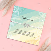 A silver heart-shaped map bracelet with a central charm, displayed on a pastel gradient card that reads 'Wanderlust' with a poem, set against a pink background with dried grass accents.