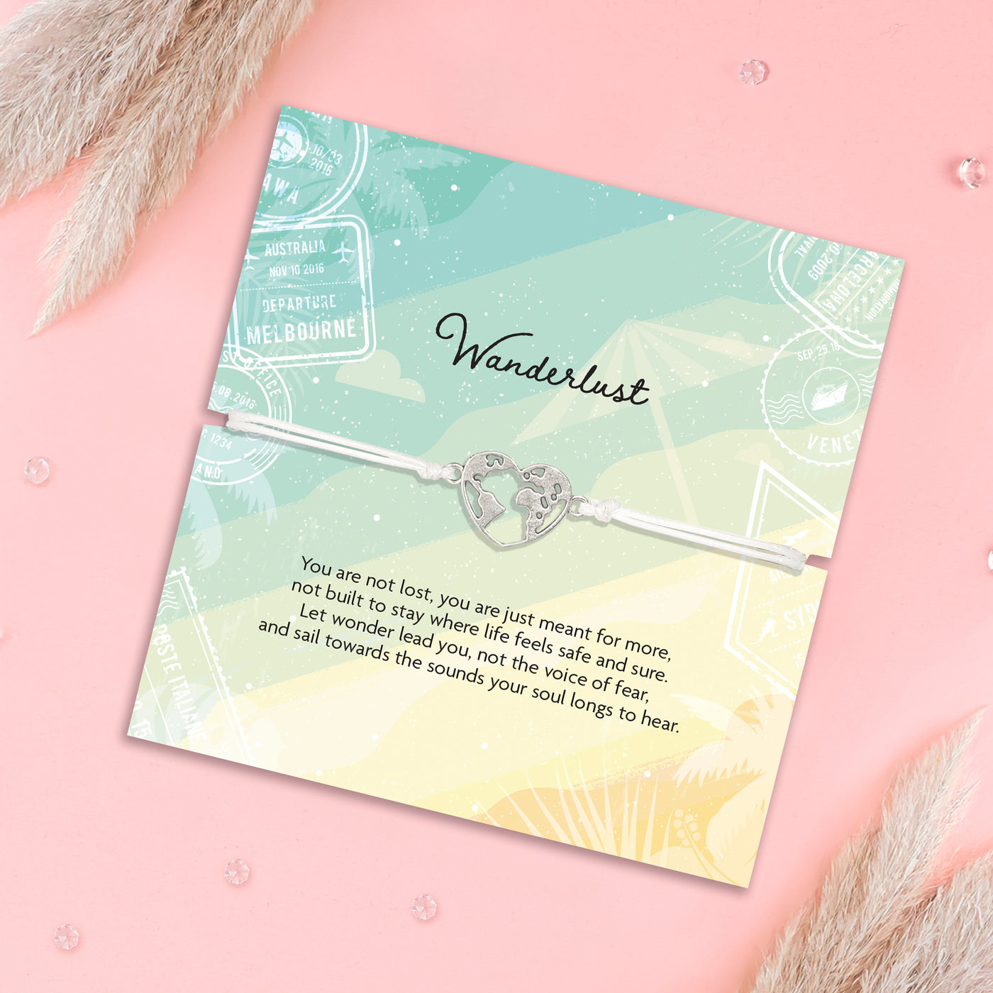 A silver heart-shaped map bracelet with a central charm, displayed on a pastel gradient card that reads 'Wanderlust' with a poem, set against a pink background with dried grass accents.