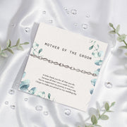 Elegant 'Mother of the Groom' card set against a silky white fabric backdrop. The card, adorned with soft blue floral designs, expresses gratitude and appreciation with a heartfelt message. Draped over the card is a sophisticated silver bracelet with intricate detailing. 
