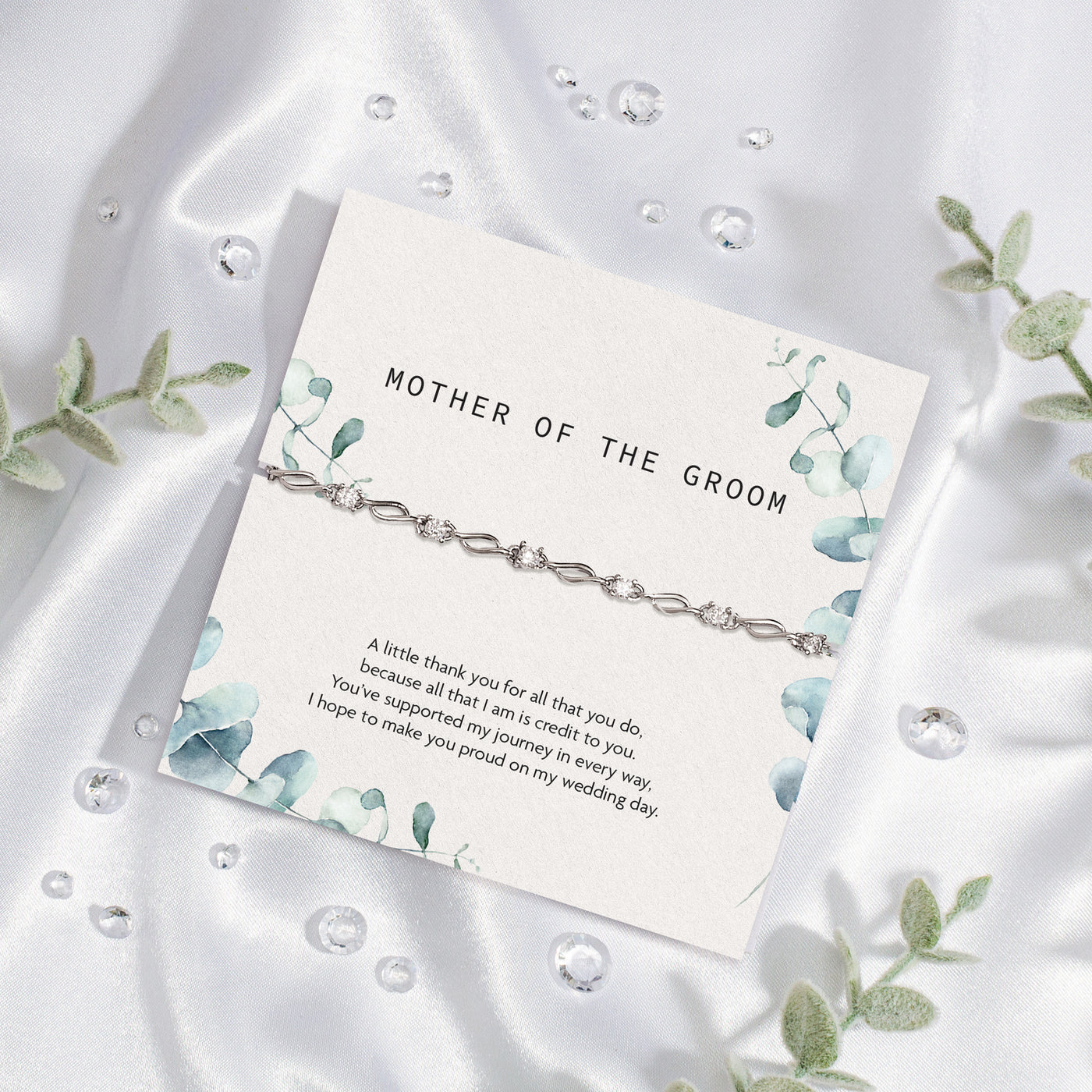 Elegant 'Mother of the Groom' card set against a silky white fabric backdrop. The card, adorned with soft blue floral designs, expresses gratitude and appreciation with a heartfelt message. Draped over the card is a sophisticated silver bracelet with intricate detailing. 