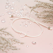 White cord friendship bracelet with a cute hollow heart on a pale pink background surrounded by gems