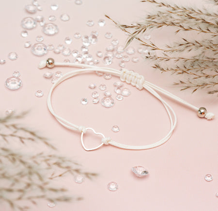 White cord friendship bracelet with a cute hollow heart on a pale pink background surrounded by gems
