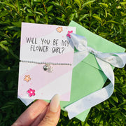 A delicate silver beaded bracelet with a small heart charm is displayed on a pink card that reads 'WILL YOU BE MY FLOWER GIRL?' A hand holds the card in front of green foliage, with a mint green gift box and white ribbon tied in a bow beside it.