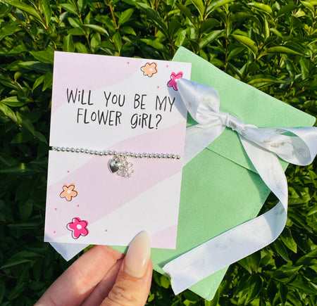 A delicate silver beaded bracelet with a small heart charm is displayed on a pink card that reads 'WILL YOU BE MY FLOWER GIRL?' A hand holds the card in front of green foliage, with a mint green gift box and white ribbon tied in a bow beside it.