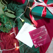 A close-up festive scene showing wrapped gift boxes, greenery, string lights, and a small jewelry card with a delicate necklace. The card reads, 'You are the real magic,' with a beautiful friendship bracelet.