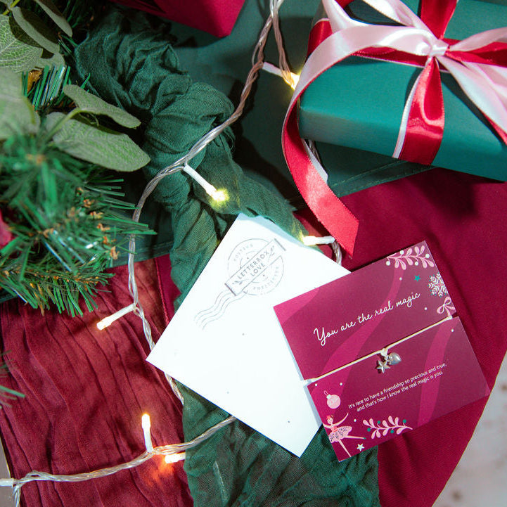 A close-up festive scene showing wrapped gift boxes, greenery, string lights, and a small jewelry card with a delicate necklace. The card reads, 'You are the real magic,' with a beautiful friendship bracelet.