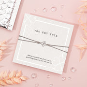"Inspirational 'You Got This' card on a soft pink background, featuring a silver four-leaf clover bracelet. The card displays subtle clover patterns and an uplifting message of belief and hope.