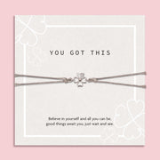"Inspirational 'You Got This' card on a soft pink background, featuring a silver four-leaf clover bracelet. The card displays subtle clover patterns and an uplifting message of belief and hope.