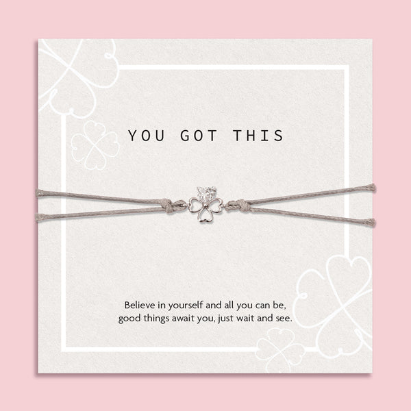 "Inspirational 'You Got This' card on a soft pink background, featuring a silver four-leaf clover bracelet. The card displays subtle clover patterns and an uplifting message of belief and hope.