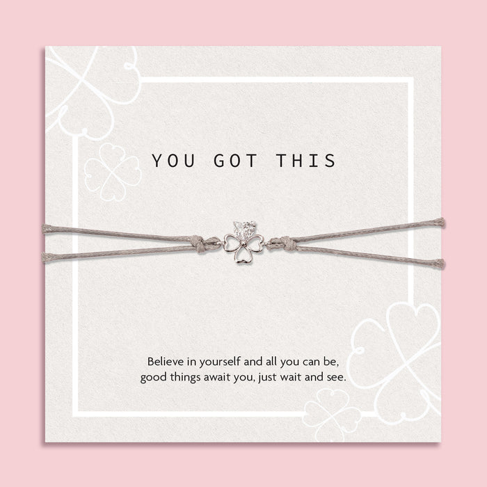 "Inspirational 'You Got This' card on a soft pink background, featuring a silver four-leaf clover bracelet. The card displays subtle clover patterns and an uplifting message of belief and hope.